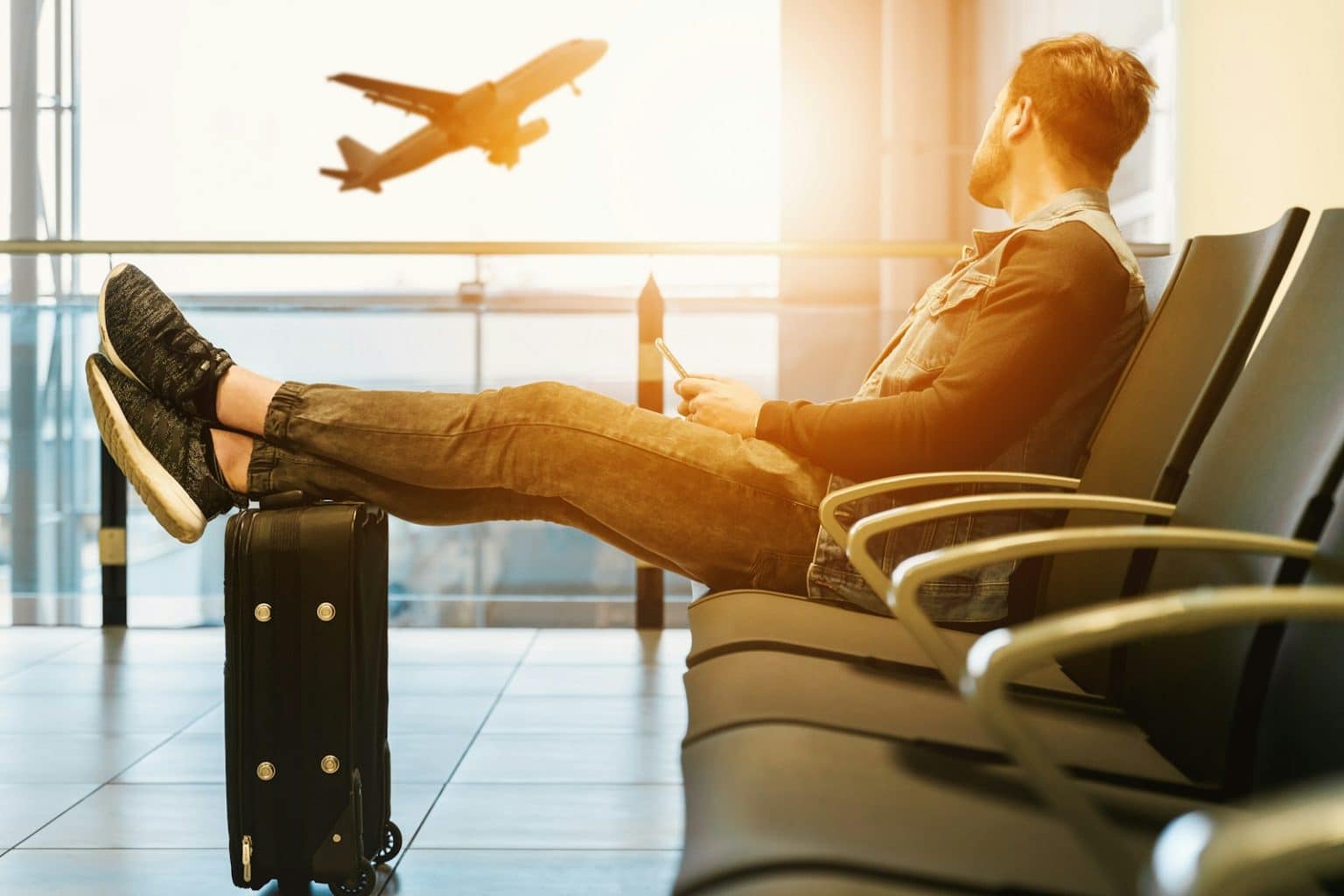 Britons Cut Back on Holiday Costs, But Risk Undervaluing Travel Insurance man sitting on gang chair with feet on luggage looking at airplane